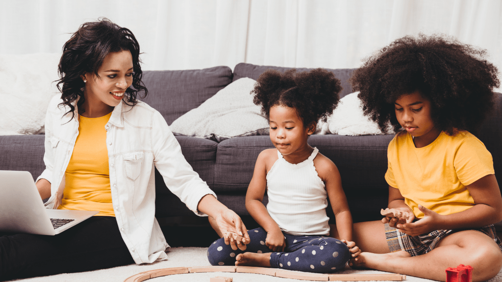 A mom working on a laptop at home while her kids play nearby, showing how to balance work and family.
